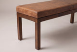 One end of the Todi bench: the square corner of the antique tan leather cushion with its stitched seam, the slim walnut-brown apron and the straight square legs below