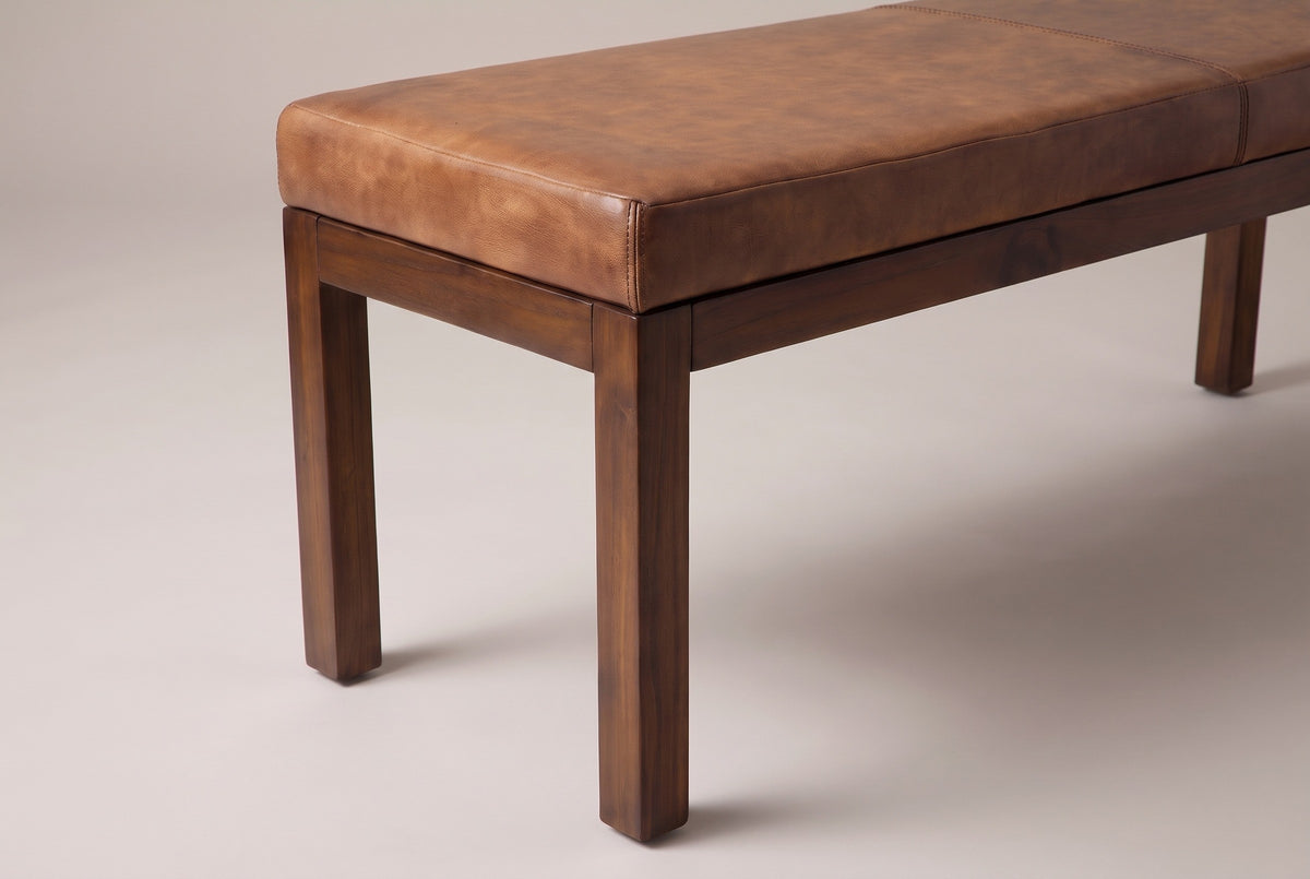 One end of the Todi bench: the square corner of the antique tan leather cushion with its stitched seam, the slim walnut-brown apron and the straight square legs below