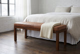 Todi antique tan leather bench at the foot of a bed with white linen bedding in a New York loft bedroom with a white-painted brick wall and a dark oak floor, a folded cream blanket on the bench