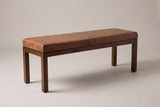 The Todi bench with its antique tan leather cushion, three-quarter view on a light studio backdrop