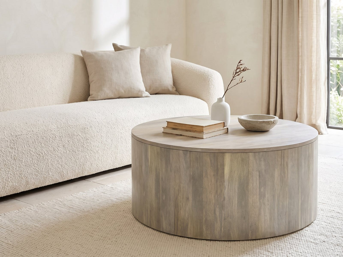Tolentino Whitewashed Mango Drum Coffee Table styled with books, a vase and a stone bowl in front of a cream sofa in a bright plaster room