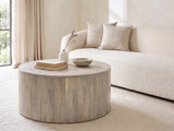 Tolentino Whitewashed Mango Drum Coffee Table seen from the side, styled with books, a vase and a stone bowl in front of a cream sofa in a bright plaster room