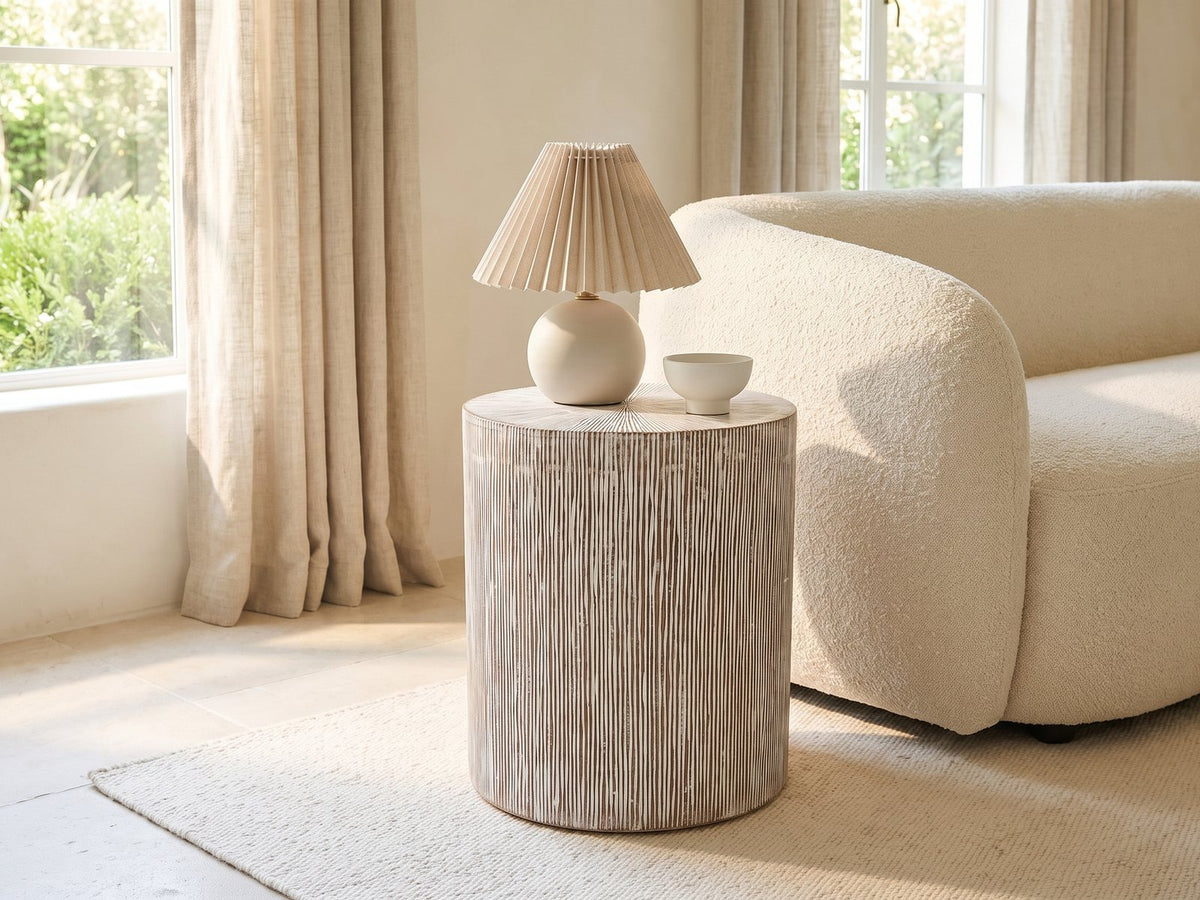 Tortora Carved Whitewash Drum Side Table with a lamp and a ceramic bowl beside a cream boucle armchair in a bright plaster room
