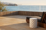 Tortorici antique white faceted concrete drum side table on a limestone terrace in Laguna Beach, a teak slat wall and a glass rail behind, a walnut lounge chair beside it (chair not included)