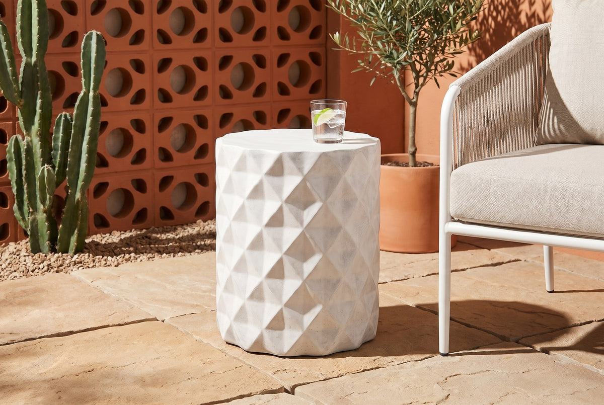 Tortorici faceted concrete side table on sand-coloured pavers in a Palm Springs patio, an orange breeze-block wall and a cactus behind, a glass of iced water on top, a white rope lounge chair beside it (chair and decor not included)