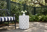 Tortorici faceted concrete side table on herringbone brick in a Savannah courtyard, a green lattice fence with jasmine and ferns behind, gardenias in a vase on top, an iron garden bench beside it (bench and decor not included)