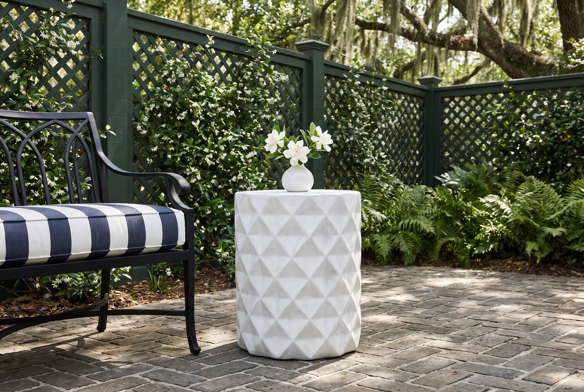 Tortorici faceted concrete side table on herringbone brick in a Savannah courtyard, a green lattice fence with jasmine and ferns behind, gardenias in a vase on top, an iron garden bench beside it (bench and decor not included)