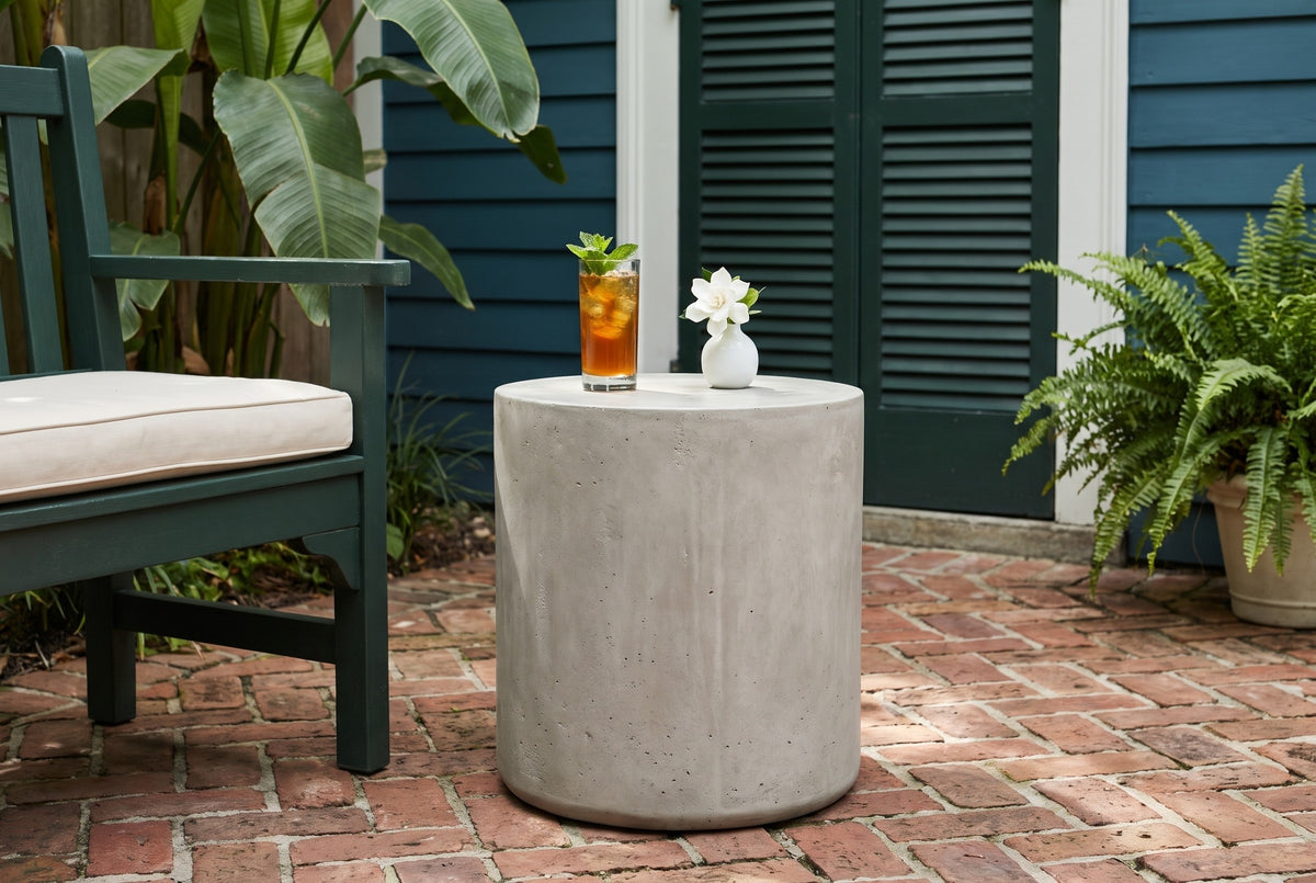 Trabia light grey concrete smooth drum side table with a glass of iced tea and a gardenia bud vase beside a green wooden armchair on a herringbone brick floor in a New Orleans Garden District courtyard, a teal clapboard wall and green shutters behind (chair and decor not included)
