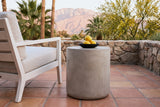 Trabia light grey concrete smooth drum side table with a black bowl of lemons beside a white wooden armchair on terracotta Saltillo tiles in Palm Springs, California at golden hour, a stacked fieldstone wall, palms and the San Jacinto mountains behind (chair and decor not included)