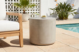 Trabia light grey concrete smooth drum side table with a glass of sparkling water and a dish of olives beside a teak armchair on shellstone pavers at a Sarasota, Florida pool terrace, a white breeze-block wall behind (chair and decor not included)