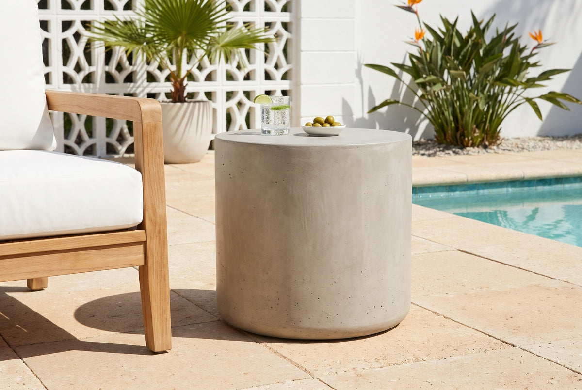 Trabia light grey concrete smooth drum side table with a glass of sparkling water and a dish of olives beside a teak armchair on shellstone pavers at a Sarasota, Florida pool terrace, a white breeze-block wall behind (chair and decor not included)