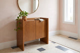 Trani oak veneer accent cabinet with a trailing plant and a round brass mirror above it, in a Charleston entryway with blush-pink plaster walls and a stone tile floor