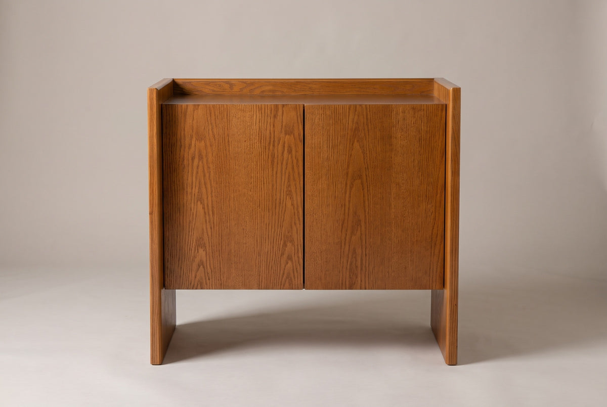 The Trani cabinet in honey oak veneer, front view on a light studio backdrop: two flat doors, a tray top and two slab ends with open floor between them
