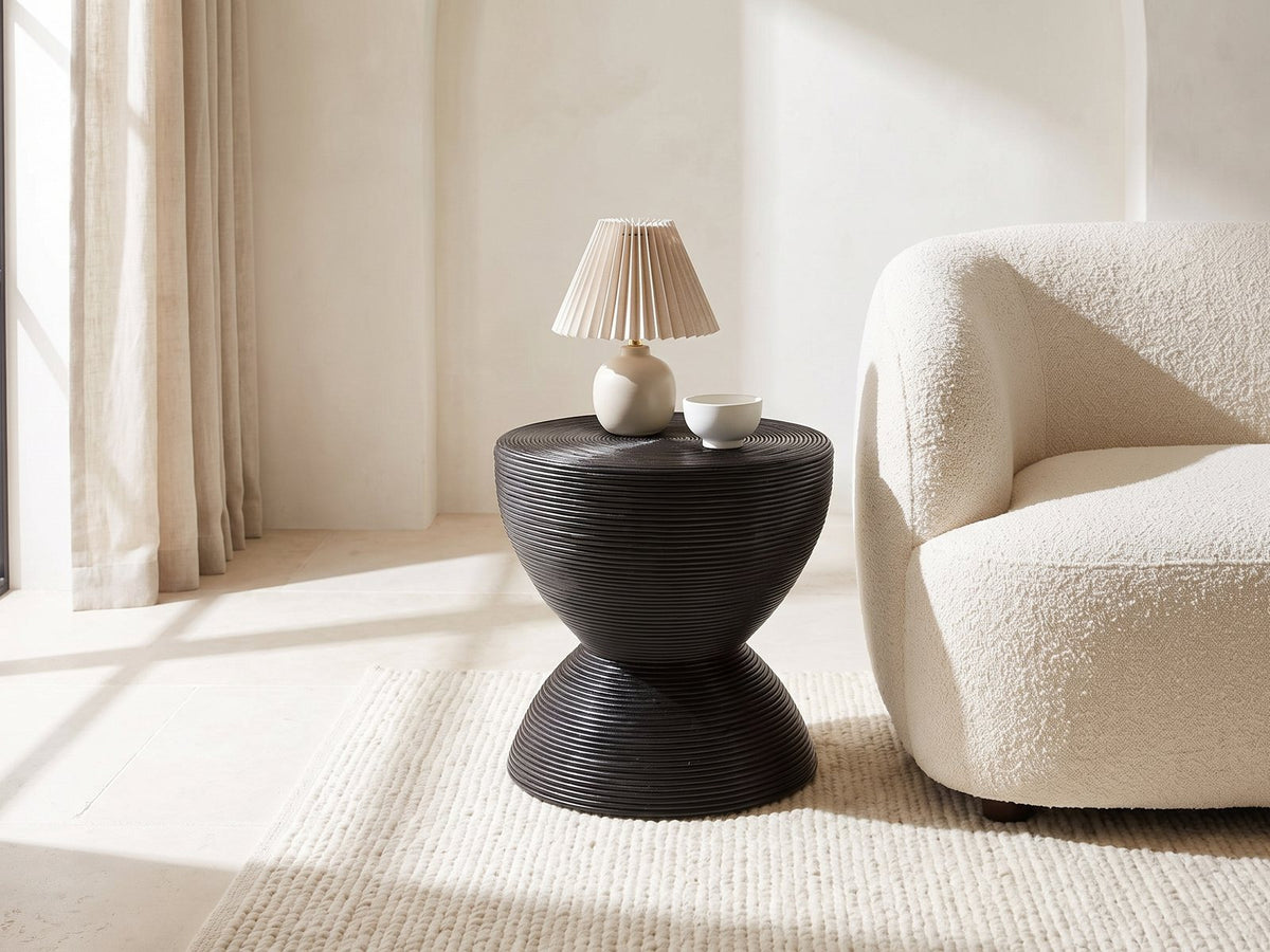 Tricase Black Rattan Hourglass Side Table with a lamp and a ceramic bowl beside a cream boucle armchair in a bright plaster room