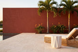 Troina antique white faceted concrete side table on travertine beside a woven lounge chair at a Naples pool terrace, a terracotta-red wall, birds of paradise and palms behind (chair not included)