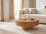 Udine Natural Wood Veneer Fluted Pedestal Coffee Table seen from the side, styled with books, a vase and a stone bowl in front of a cream sofa in a bright plaster room