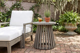 Valderice slatted acacia side table on a brick courtyard floor in Savannah, Georgia, an old rose-grey brick wall, ferns and Spanish moss behind, a white-cushioned lounge chair beside it (chair, glass and plant not included)