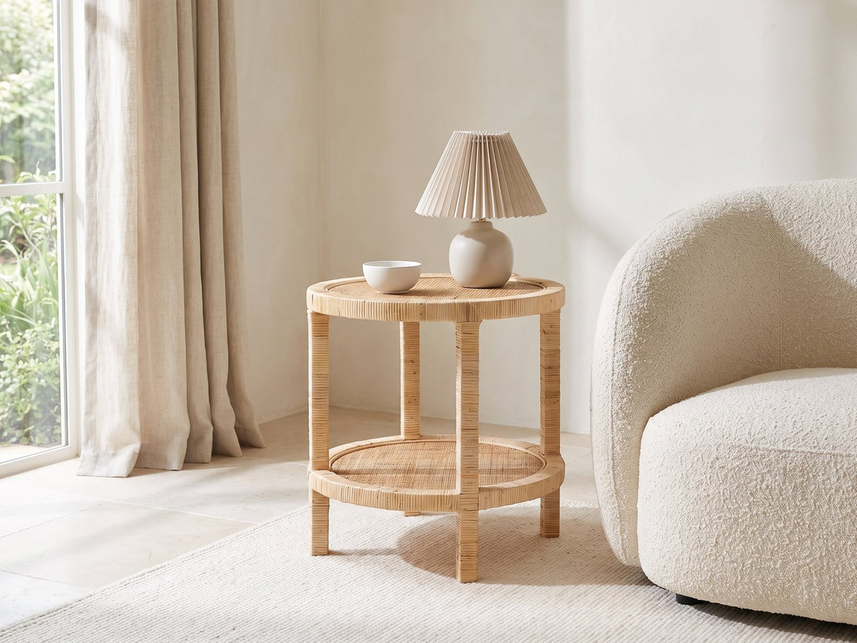 Valsinni Rattan Round Two-Tier Side Table with a lamp and a ceramic bowl beside a cream boucle armchair in a bright plaster room