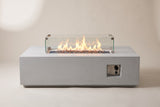The Vulcano fire pit table seen from the front: the long concrete body, the glass wind guard with a lit flame and the control niche at the right-hand end