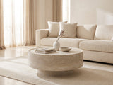 Zagarolo Faux Travertine Round Drum Coffee Table styled with books, a vase and a stone bowl in front of a cream sofa in a bright plaster room