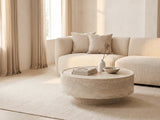 Zagarolo Faux Travertine Round Drum Coffee Table seen from the side, styled with books, a vase and a stone bowl in front of a cream sofa in a bright plaster room