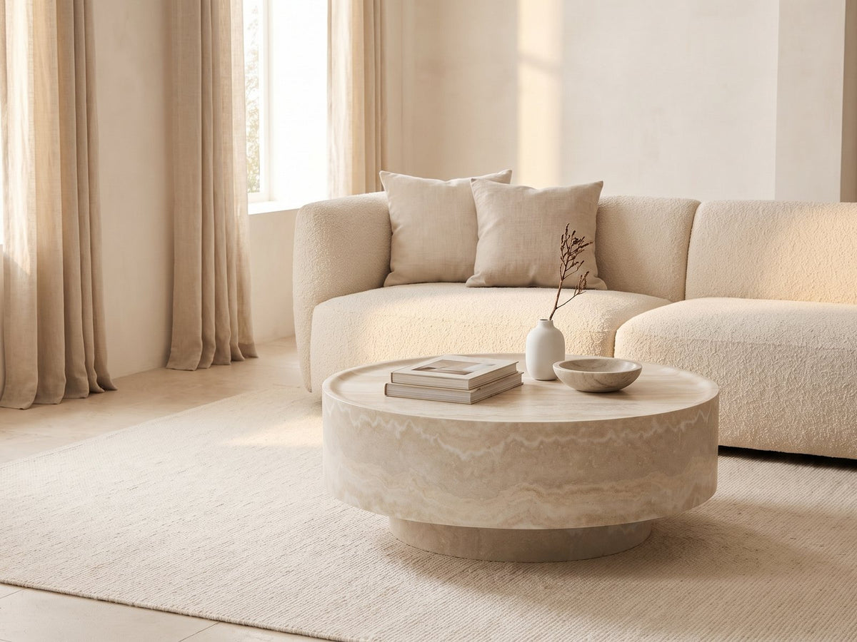 Zagarolo Faux Travertine Round Drum Coffee Table seen from the side, styled with books, a vase and a stone bowl in front of a cream sofa in a bright plaster room