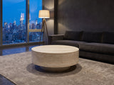 Zagarolo Faux Travertine Round Drum Coffee Table on a wool rug in front of a velvet sofa in a city penthouse at blue hour