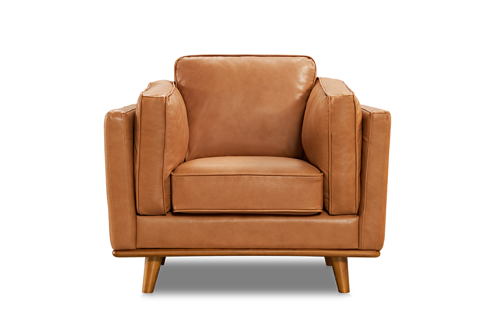 A Valencia Artisan Leather Accent Chair in Cognac color, placed on a rug in a room with wooden floors. It's beside a floor lamp, near a large window showing a snowy outdoor view. An abstract painting hangs on the wall above it, and a potted plant is nearby, highlighting the chair's stylish and cozy appeal.
