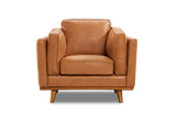 A Valencia Artisan Leather Accent Chair in Cognac color, placed on a rug in a room with wooden floors. It's beside a floor lamp, near a large window showing a snowy outdoor view. An abstract painting hangs on the wall above it, and a potted plant is nearby, highlighting the chair's stylish and cozy appeal.
